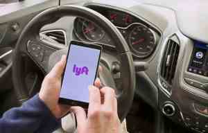A person sits in the driver’s seat holding a phone with the Lyft app opening.