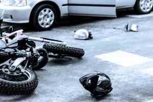 A crashed motorcycle lies near a damaged car. A helmet and broken side-view mirror lie on the ground as well.
