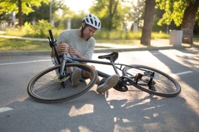 Does Health Insurance Cover Bicycle Accident Injuries? A man wearing a helmet while sitting on the asphalt of a city street after a bicycle crash, his leg injured, as he wonders, “Does health insurance cover bicycle accident injuries?