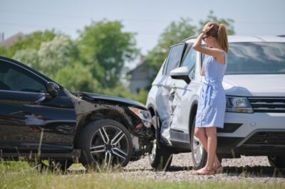 A woman with a hand on her head stands next to a damaged black car and a damaged white car, talking on her phone, wondering if she should get a lawyer for her car accident.